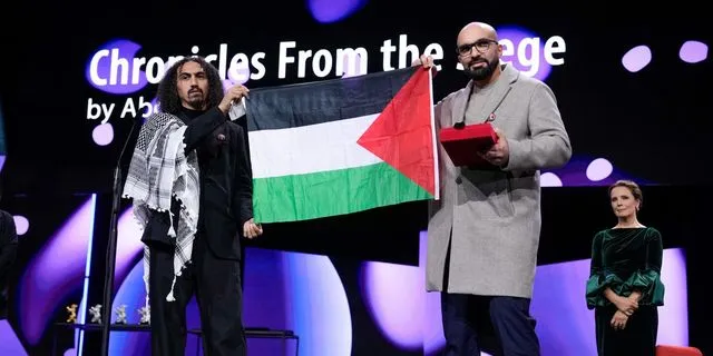 Ο σκηνοθέτης Αμπντάλα Αλ-Χατίμπ και ο Τακιγιεντίν Ισάαντ κρατούν παλαιστινιακή σημαία μετά την απονομή του βραβείου GWFF για την καλύτερη πρώτη μεγάλου μήκους ταινία στο Chronicles from the Siege, κατά την τελετή απονομής του Διεθνούς Φεστιβάλ Κινηματογράφου Μπερλινάλε το Σάββατο 21 Φεβρουαρίου 2026