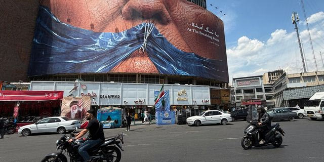 Οχήματα περνούν δίπλα από μια πινακίδα που απεικονίζει το Στενό του Ορμούζ και ραμμένα χείλη του Προέδρου των ΗΠΑ Ντόναλντ Τραμπ σε μια πλατεία στο κέντρο της Τεχεράνης