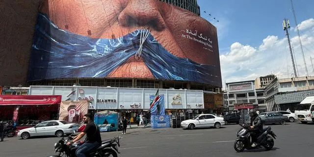 Οχήματα περνούν μπροστά από μια διαφημιστική πινακίδα με γραφικό που απεικονίζει τα Στενά του Ορμούζ και τα ραμμένα χείλη του Ντόναλντ Τραμπ, σε πλατεία στο κέντρο της Τεχεράνης, στο Ιράν